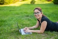 Happy red-haired girl in glasses lies on the lawn in the park and types on the laptop keyboard. Young caucasian female