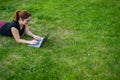 Happy red-haired girl in glasses lies on the lawn in the park and types on the laptop keyboard. Young caucasian female