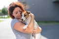 Happy red-haired woman hugs her little dog for a walk outdoors. Jack Russell Terrier licks the face of the female owner.