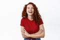 Happy redhead girl with curly hair, laughing positive with eyes closed, cross arms on chest and chuckling carefree