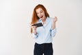 Happy redhead girl winning in mobile phone video game, saying yes and smiling, celebrating online victory, standing over Royalty Free Stock Photo