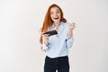 Happy redhead girl winning in mobile phone video game, saying yes and smiling, celebrating online victory, standing over Royalty Free Stock Photo