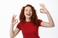 Happy redhead woman feeling carefree, showing okay signs and laughing cheerful with closed eyes, enjoy life, say alright