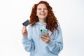 Happy redhead woman rejoicing, holding smartphone and plastic credit card, close eyes and laugh from overjoy, standing Royalty Free Stock Photo