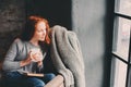 Happy redhead woman relaxing at home in cozy winter or autumn weekend with book and cup of hot tea, sitting in soft chair Royalty Free Stock Photo