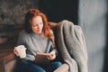 Happy redhead woman relaxing at home in cozy winter or autumn weekend with book and cup of hot tea, sitting in soft chair Royalty Free Stock Photo