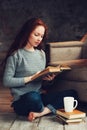 Happy redhead woman relaxing at home in cozy winter or autumn weekend with book and cup of hot tea, sitting in soft chair