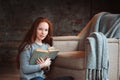 Happy redhead woman relaxing at home in cozy winter or autumn weekend with book and cup of hot tea, sitting in soft chair Royalty Free Stock Photo