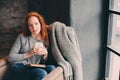 Happy redhead woman relaxing at home in cozy winter or autumn weekend with book and cup of hot tea, sitting in soft chair