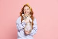 Happy redhead young girl with long curly hair holding cute little puppy of corgi dog isolated on pink background