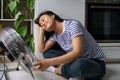 Happy relaxed Asian girl rejoices in flow of cold air from fan sits on floor in kitchen of apartment Royalty Free Stock Photo