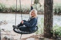 Happy, relaxed blonde woman sits on a tree swing near a river, looking happy