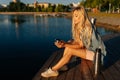 Happy relaxed blonde young woman holding cup with hot coffee from thermos sitting on beach by wooden pier on sunny Royalty Free Stock Photo