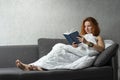 Happy resting woman. Young ginger girl on the sofa under blanket reads book