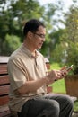 A happy retired Asian man using his smartphone, texting someone while sitting on a bench in a park Royalty Free Stock Photo
