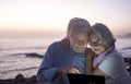 Happy retired couple having fun sitting on the cliffs, dusk light looking at the same tablet. Happy retirement concept. Sea in the Royalty Free Stock Photo