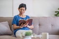 A happy retired 60s Asian woman reading a book while having her coffee on a sofa at home. Royalty Free Stock Photo