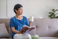 A happy retired 60s Asian woman reading a book while having her coffee on a sofa at home. Royalty Free Stock Photo