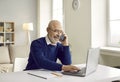 Happy senior man sitting at his desk, talking on his mobile phone and using his laptop Royalty Free Stock Photo