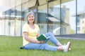 Happy retired woman sitting on the grass resting on a summer day and reading a book, looking at the camera and smiling Royalty Free Stock Photo