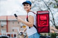 Happy rider woman wearing red backpack delivering food on a bike, checking order with smart phone while standing on street in city Royalty Free Stock Photo