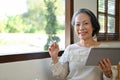 Happy 60s aged Asian woman sitting by the window in her living room and using tablet Royalty Free Stock Photo