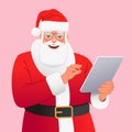 Happy Santa Claus in glasses and a red hat stands with a tablet in his hand. Cartoon Santa makes a list of gifts on a personal Royalty Free Stock Photo