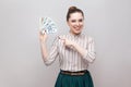 Happy satisfied attractive young businesswoman in striped shirt standing, holding many dollars, pointing finger and looking at