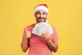 Happy satisfied bearded man in red t-shirt and santa claus hat showing thumbs up holding fan of dollars, looking at camera with Royalty Free Stock Photo