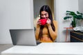 Happy satisfied young woman enjoying cup of coffee at home while using laptop smiling girl drinking tea or hot chocolate while Royalty Free Stock Photo