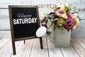 Happy Saturday text on blackboard easel with flower bouquet on wooden background