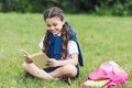 happy schoolgirl reading book while sitting on grass