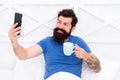 Happy selfie. Good morning. Bearded man using mobile technology in bed. Handsome guy talking phone and drinking coffee Royalty Free Stock Photo