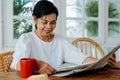 Happy senior Asian female in casual clothes reading interesting article in newspaper while having breakfast at home Royalty Free Stock Photo