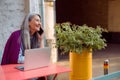 Happy senior Asian woman talks on phone sitting at table with laptop