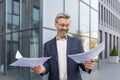 Happy senior businessman standing on the street near the modern office holding papers in his hands, satisfied with the Royalty Free Stock Photo