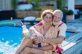 Happy senior couple enjoying drinks when relaxing and sitting by swimming pool in summer, looking at camera. Royalty Free Stock Photo