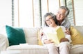 Happy senior couple are sitting on the couch together and reading a book and looking sideways in living room at home Royalty Free Stock Photo