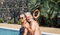 Happy senior couple on vacation in swimwear sitting at the swimming pool - man with beard and white hair hugs his wife laughing. Royalty Free Stock Photo