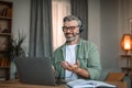 Happy senior european man with beard in headset and glasses gesticulates and looks at laptop