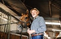 A senior man with a hat standing close to a horse in a stable, holding it. Royalty Free Stock Photo