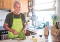 Happy senior man with white beard and hair in the kitchen while cuts and cleans the broccoli to prepare an healthy soup Royalty Free Stock Photo