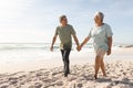 Happy senior multiracial couple holding hands while walking on sand at sunny beach against sky Royalty Free Stock Photo