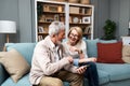 Happy senior old couple holding smartphone looking at cellphone screen laughing relaxing sit on sofa together, smiling elder