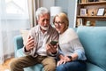 Happy senior old couple holding smartphone looking at cellphone screen laughing relaxing sit on sofa together, smiling elder