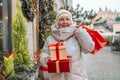 Happy senior 50s blonde woman having fun holding shopping bag full of Christmas gift boxes on city street. Seasonal Royalty Free Stock Photo