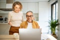 Happy senior spouses man and woman sitting at kitchen table in front of laptop computer, looking at screen and smiling Royalty Free Stock Photo