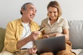 Happy senior spouses shopping online on laptop, man giving credit card to his wife, sitting on sofa at home Royalty Free Stock Photo