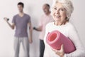 Senior woman holding pink joga mat, men exercising in background