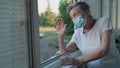 Happy senior woman in mask waves to family from window hospital. Masked elderly female glad that children came visit her Royalty Free Stock Photo